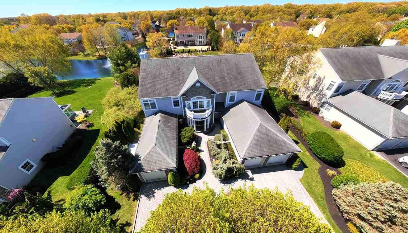 Aerial daytime view of 38 Hamlet Woods Drive showing the gray shingle Colonial, paver motor court, and the community lake at left
